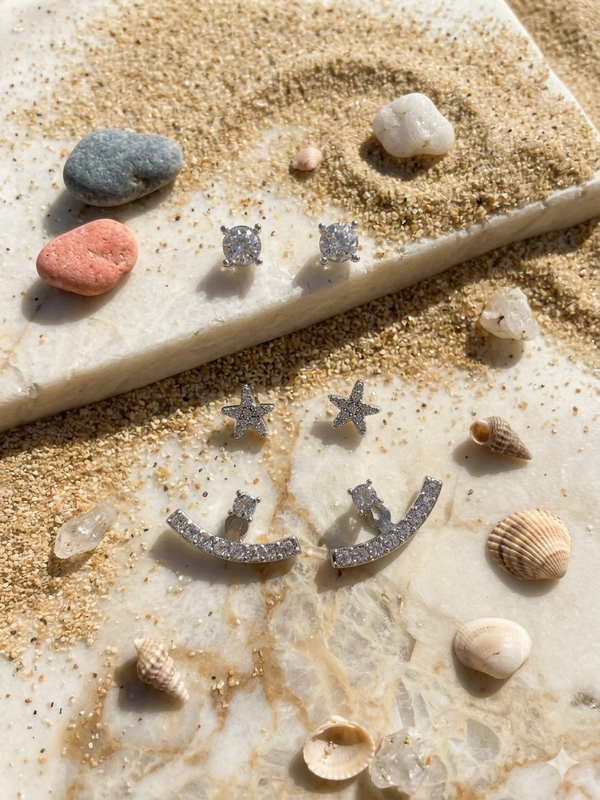 Silver star and smiley face earrings on a sandy surface with shells and stones.