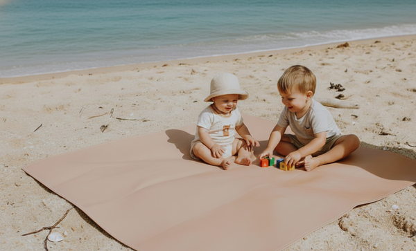 Two children playing on a beach with a pink blanket and toys.