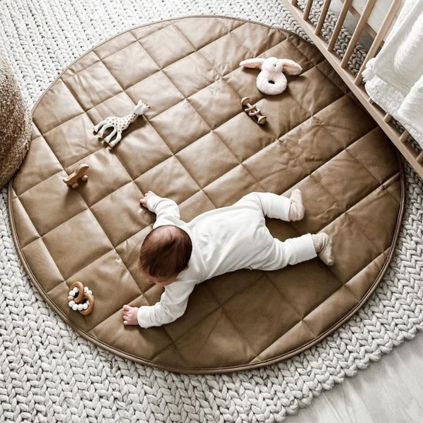 A baby lying on a round quilted floor mat in tan and gray colors with plush toys attached to it.