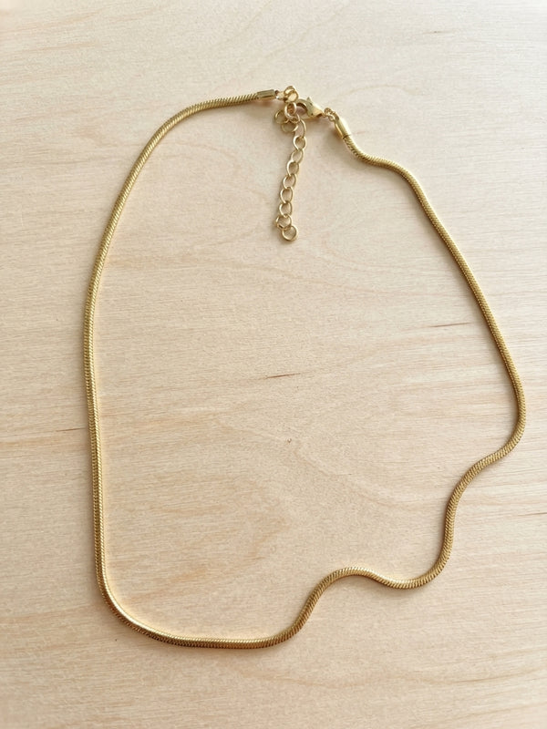 A gold snake chain necklace with an adjustable lobster clasp and extender chain, displayed on a light wood grain surface.