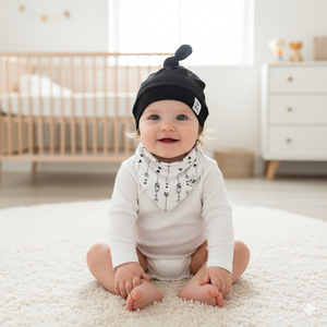 Baby sitting on a rug in a nursery wearing a black hat and white onesie with a bandana.